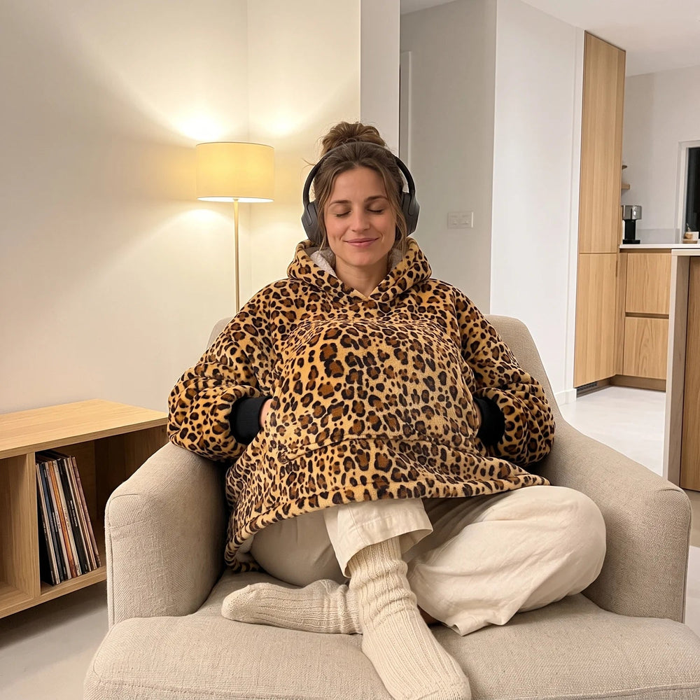 Woman in the Golden Leopard blanket hoodie listening to music in an armchair, knees tucked in