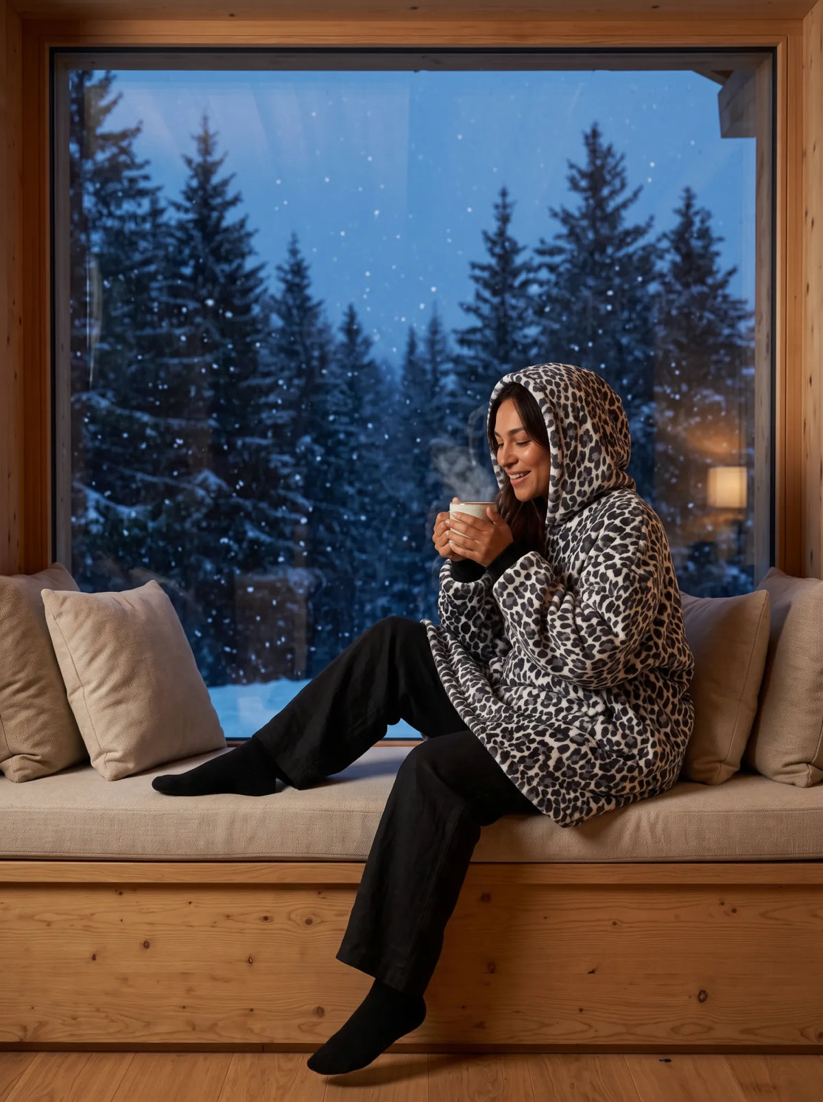 Woman wearing the Snow Leopard blanket hoodie on a window bench in a mountain chalet with a hot chocolate while snow falls outside
